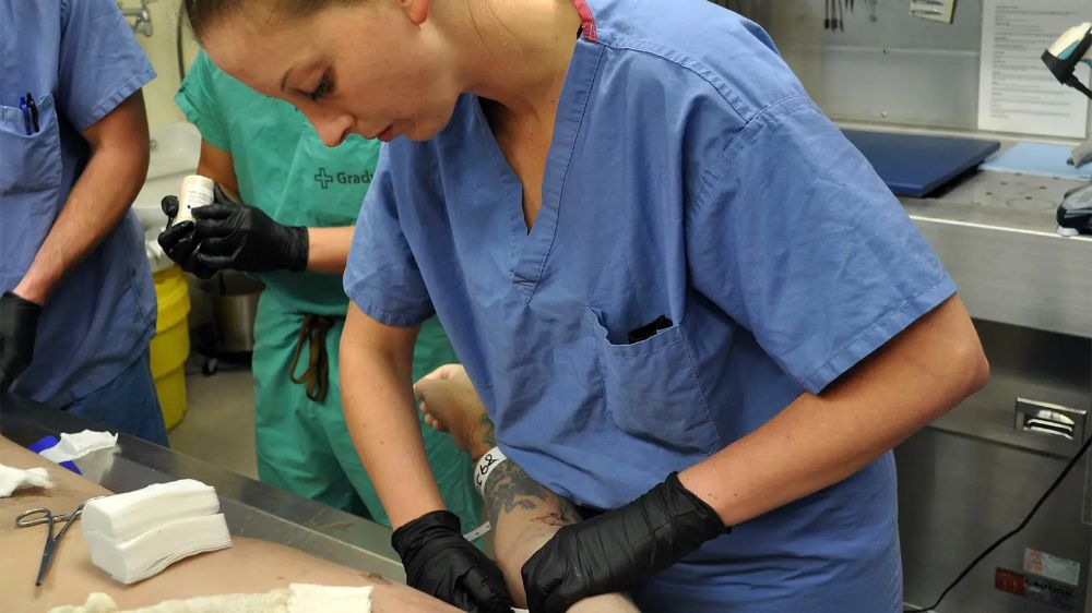 A person wearing blue medical scrubs and black gloves is preparing or treating a tattooed arm in a clinical or medical setting, with another person in green scrubs and medical tools visible nearby.