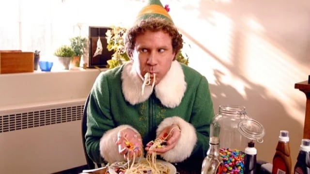 A man dressed as an elf in a green suit and hat eats spaghetti with his hands, surrounded by candy, syrup bottles, and a large jar of sweets on the table. He looks off to the side with a playful expression.