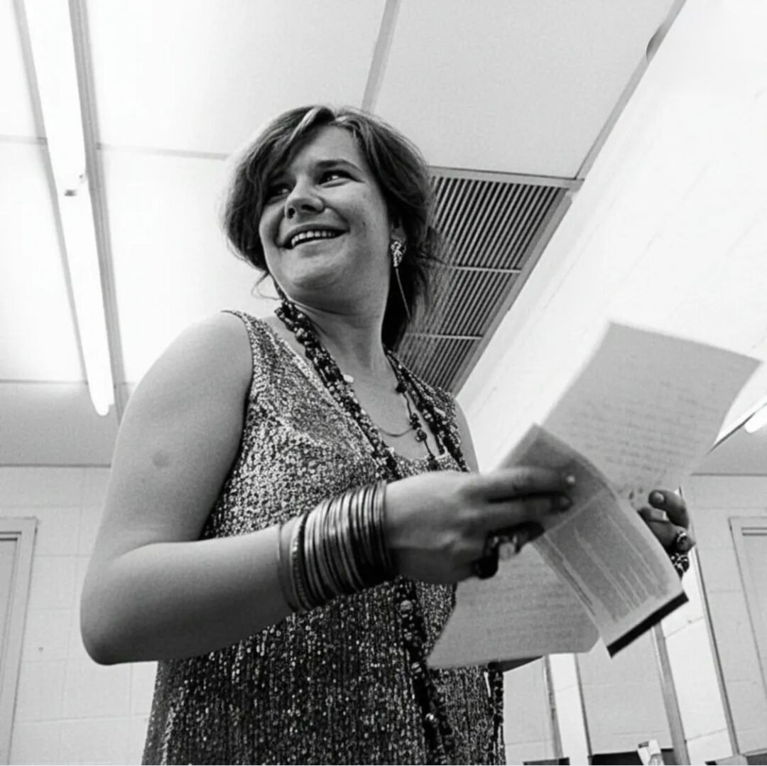 A woman in a sparkly sleeveless top smiles while holding papers in both hands. She wears multiple bangles and beaded necklaces, standing in a brightly lit room with a tiled wall and ceiling lights.