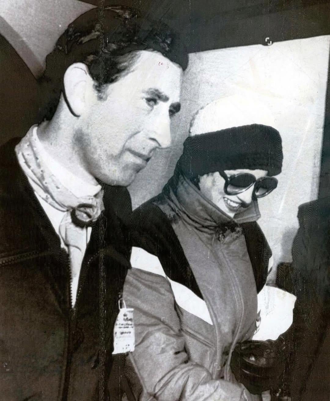 A man and a woman dressed in winter clothing stand close together. The man wears a scarf and jacket, and the woman, smiling, is in a beanie, sunglasses, and a ski jacket. The photo appears to be black and white.