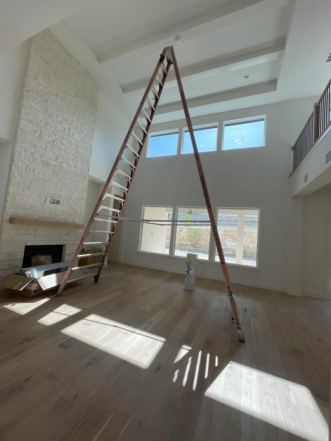 A tall ladder is set up in the center of a spacious, sunlit living room with high ceilings, large windows, light wood floors, and a stone fireplace. Shadows from the ladder fall across the floor.