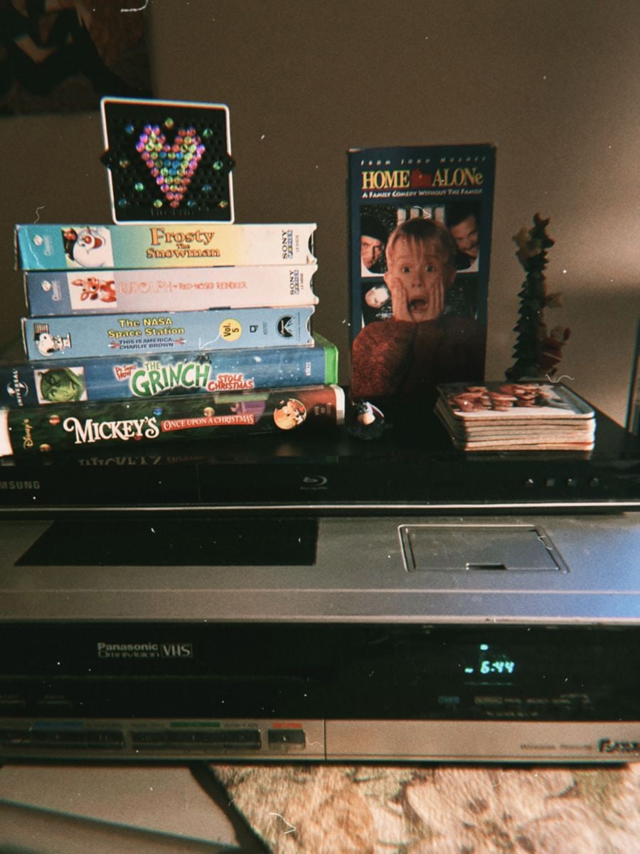 A stack of VHS tapes, including "Home Alone," sits on a VCR. The tapes feature holiday and cartoon themes. Nearby are coasters, a small decorated tree, and a heart-shaped bead art piece.