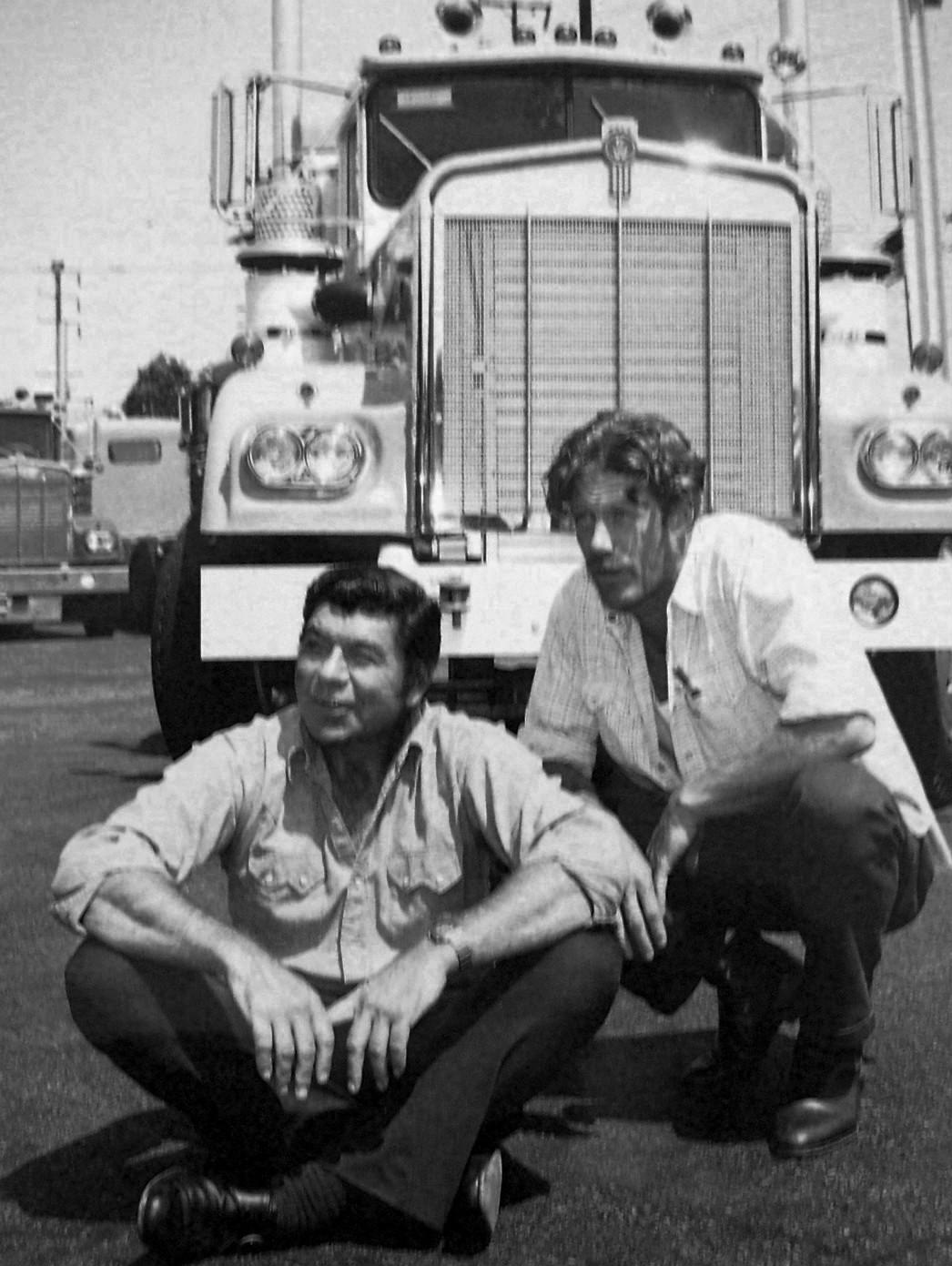 Two men in casual shirts sit and crouch on the pavement in front of a large semi-truck, likely in a parking lot, with another truck visible in the background. The image is in black and white.