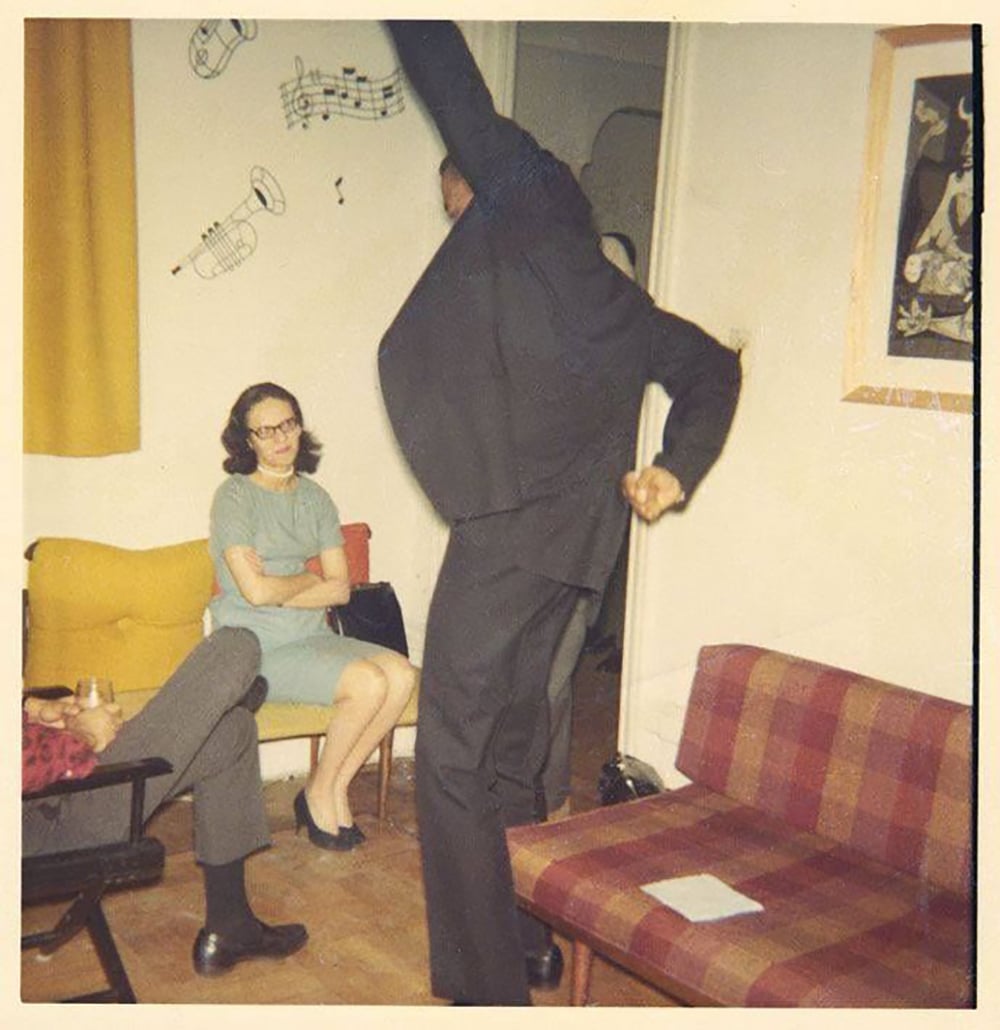 A man in a suit dances energetically in a living room while two women sitting on a yellow couch watch him. Musical notes and instruments are drawn on the wall behind them.