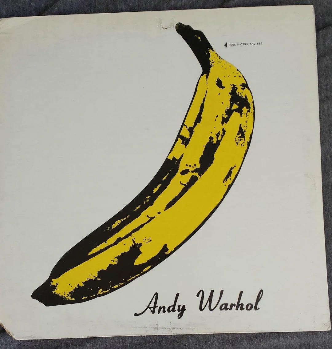 A yellow banana with black spots is illustrated on a white background, with "Andy Warhol" written in cursive at the bottom. The top right corner has the small text, "PEEL SLOWLY AND SEE.