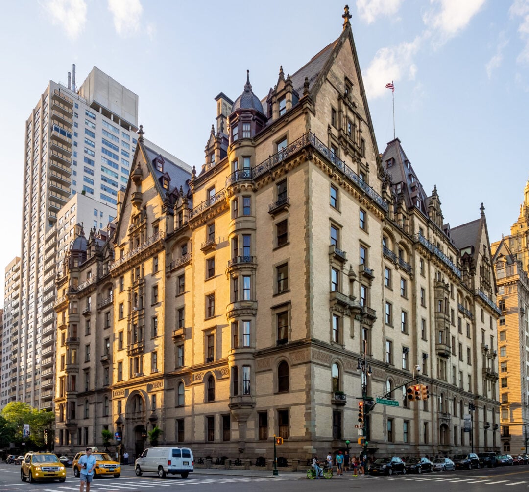 A historic, ornate stone building with peaked roofs and decorative details stands on a city corner, surrounded by modern high-rises, cars, and yellow taxis on a sunny day.