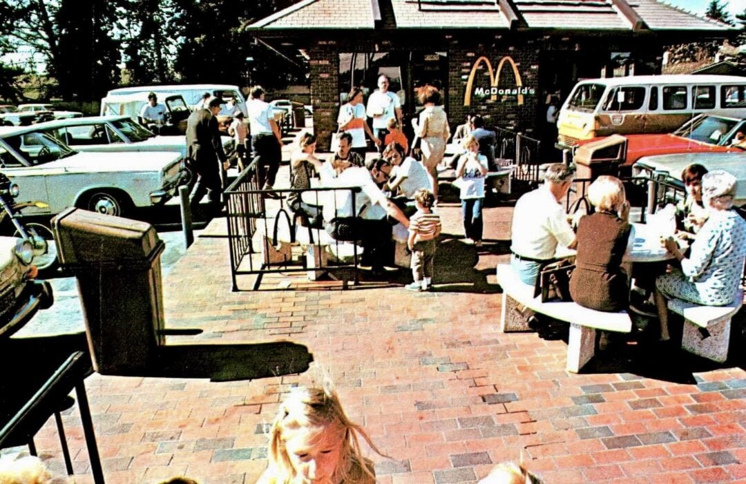 People sit and stand outside a vintage McDonald's, with classic cars parked nearby. Adults and children eat at outdoor tables on a sunny day, creating a busy, lively atmosphere.
