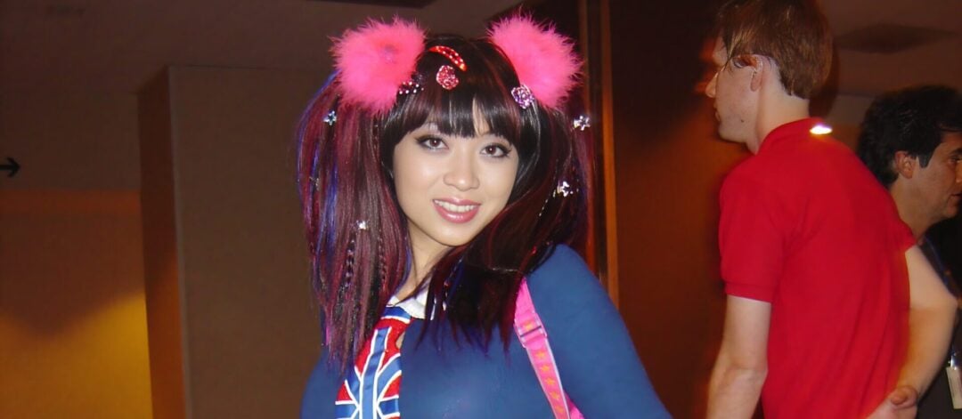 A woman wearing a colorful cosplay outfit with UK flag designs, pink accessories, white boots with red hearts, and pink furry hairbands poses in a hallway with patterned carpet. Other people are in the background.