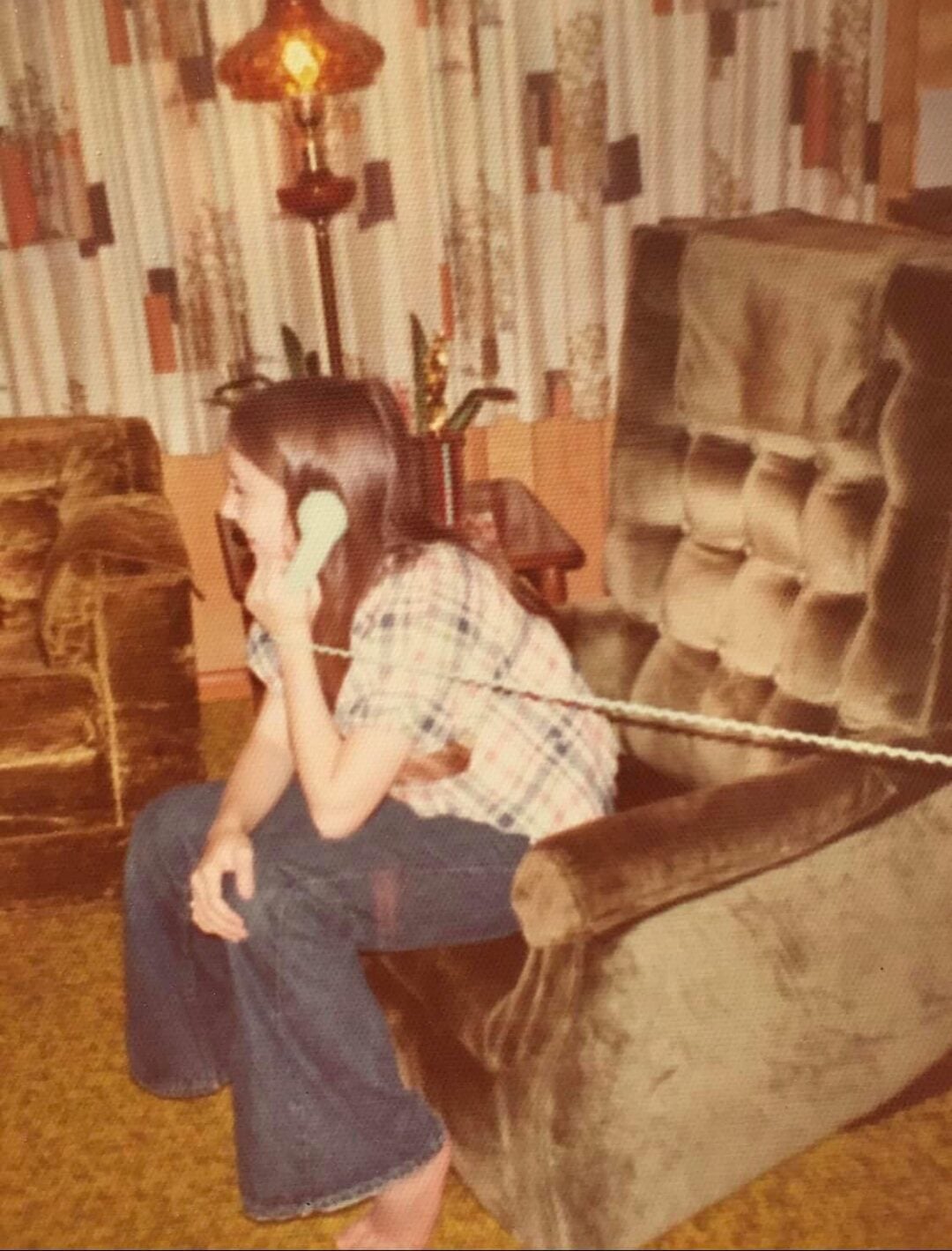A young woman sits in a vintage living room on a green velvet armchair, talking on a corded phone. She wears a plaid shirt and flared jeans. Retro decor, including patterned curtains and a lamp, is visible in the background.