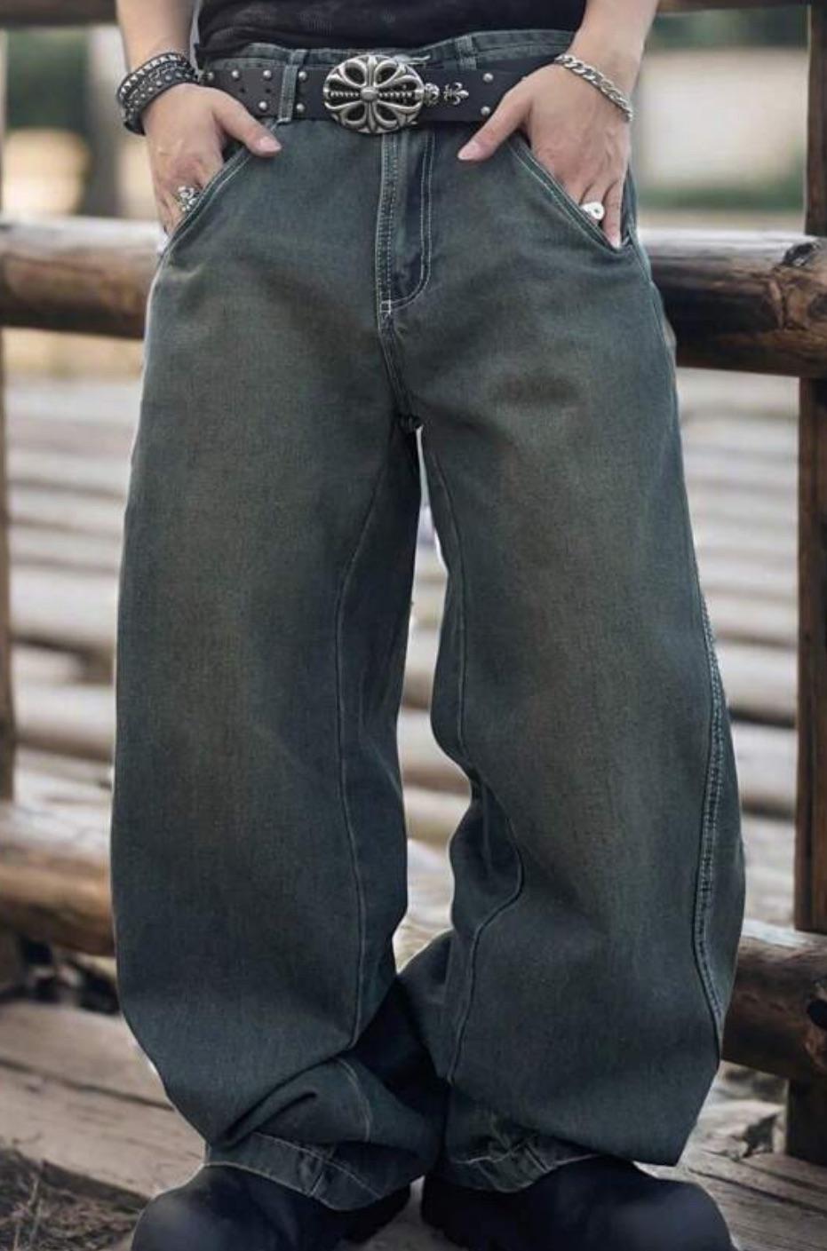 Person standing outdoors wearing baggy jeans, black shoes, a studded belt with a large ornate buckle, rings, and bracelets, with hands in pockets. Wooden railing and blurred background visible.