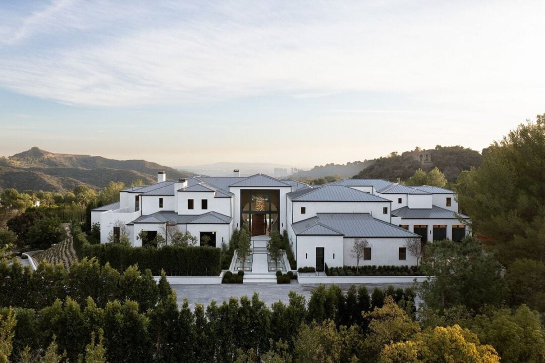 A large, modern white mansion with multiple sections and a metal roof, surrounded by trees and hedges, set against a backdrop of rolling hills under a partly cloudy sky.