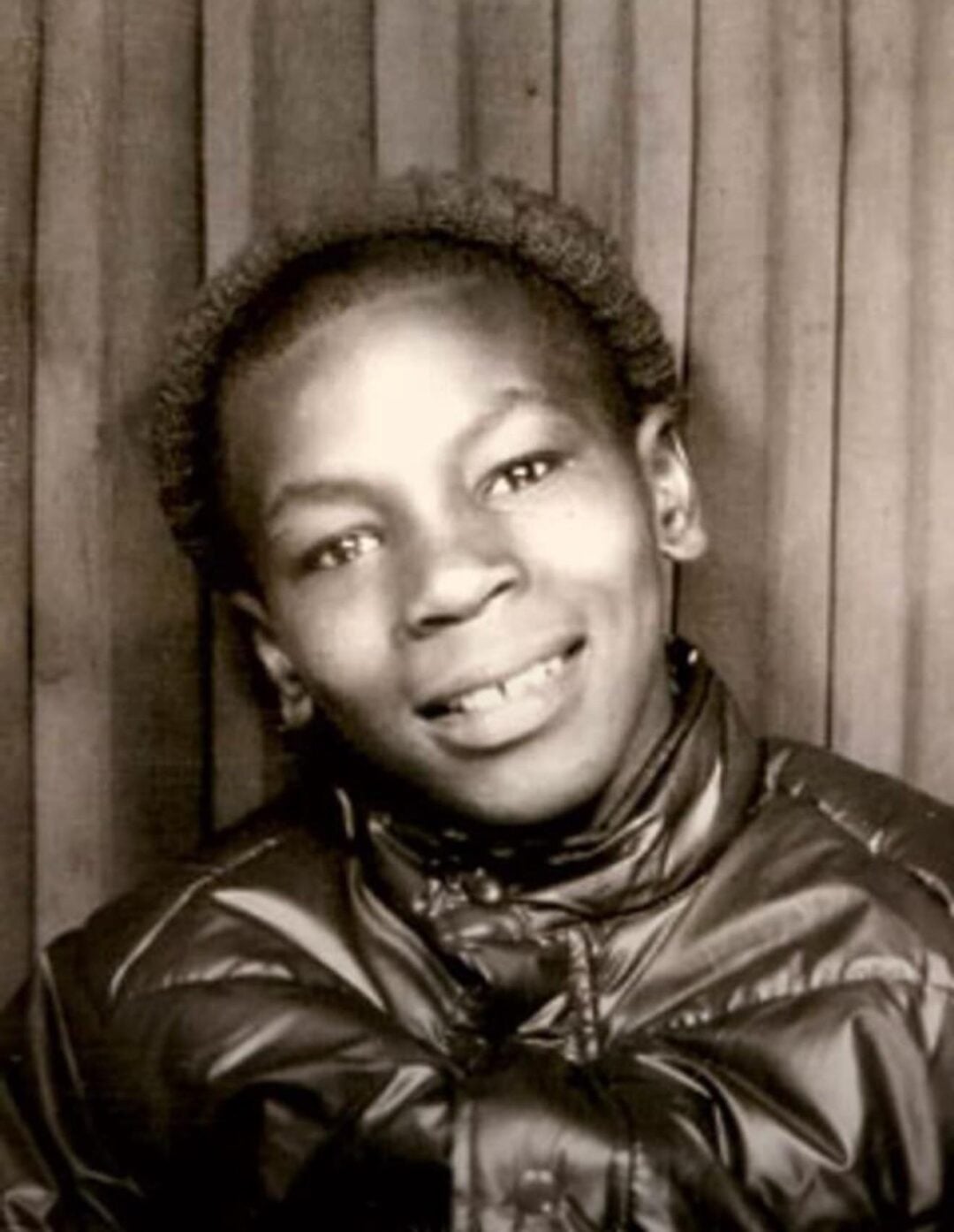 Black-and-white photo of a young boy smiling, wearing a shiny, zippered jacket, standing in front of a vertical wooden panel background.