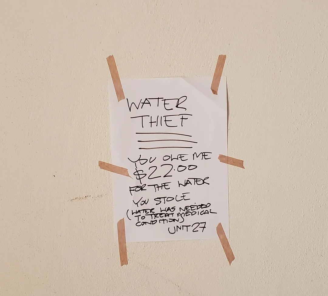 A handwritten sign taped to a wall reads: "WATER THIEF. You owe me $22.00 for the water you stole (Water has needed to treat medical condition). UNIT 27" with emphasis on "water thief.