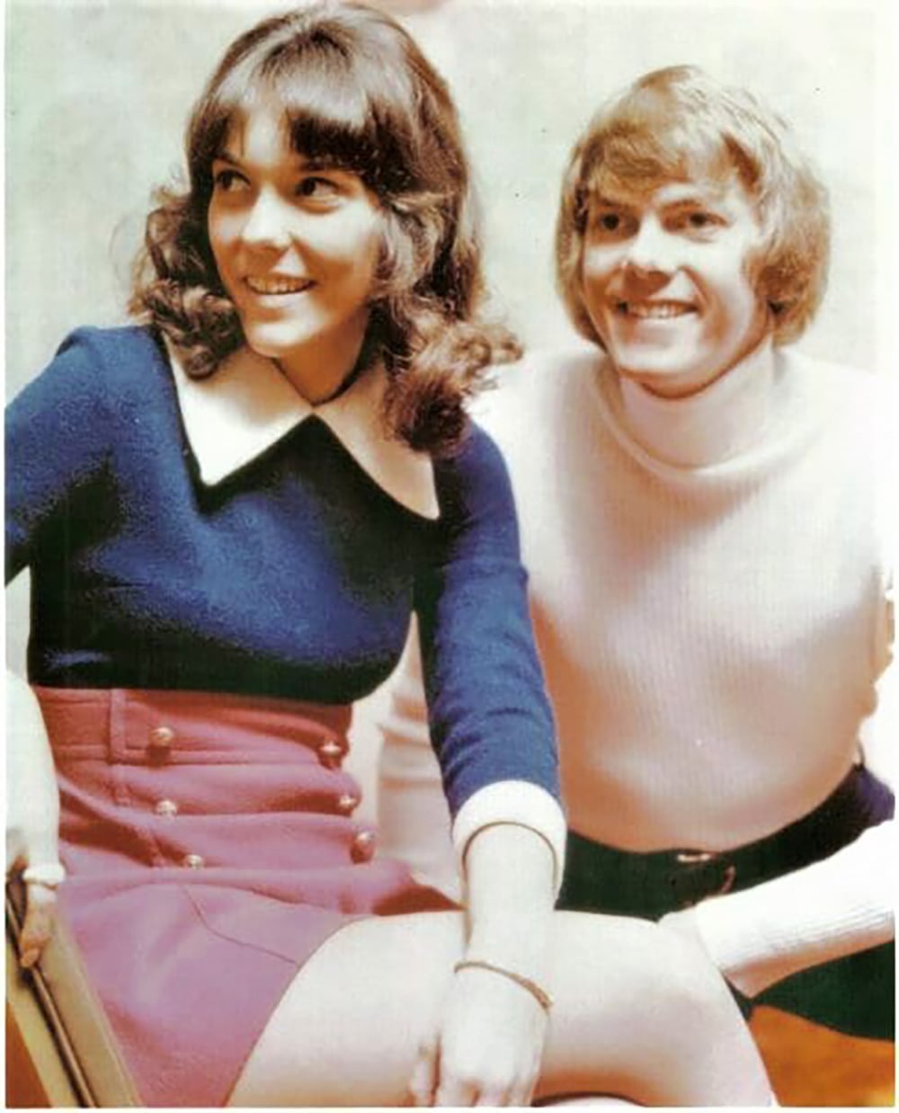 A smiling woman with brown hair in a blue top and pink skirt sits next to a smiling man with blond hair in a white turtleneck sweater. They both look slightly off-camera in a bright, softly-lit setting.