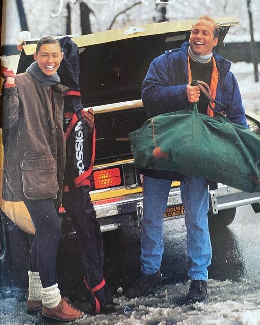 A smiling man and woman stand by an open car trunk in winter. The man holds a large green duffel bag, and the woman holds winter gear. Snow is on the ground, and both are dressed warmly in coats and boots.