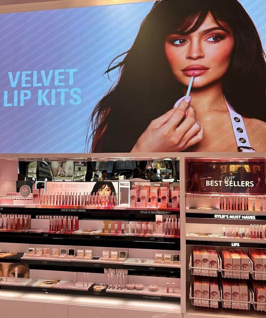 A Kylie Cosmetics display in a store features shelves of makeup products and a large screen showing a woman applying lip gloss with the text "VELVET LIP KITS." A banner announces its launch at Mumbai Duty Free.