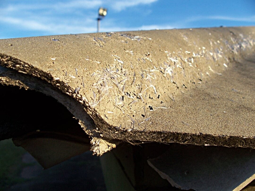 Close-up of a damaged, fraying sheet of asbestos cement, showing its rough texture and fibrous material with a blurred outdoor background.