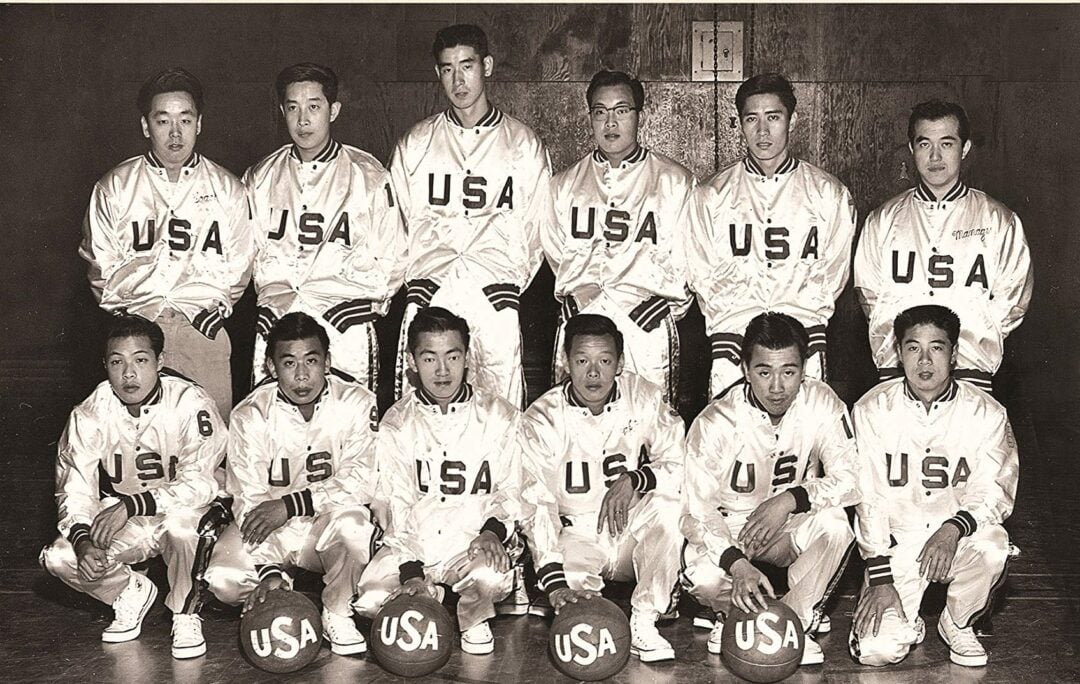 A vintage black-and-white photo of a men's basketball team wearing matching USA jackets, posed in two rows indoors. The front row kneels with basketballs, while the back row stands.