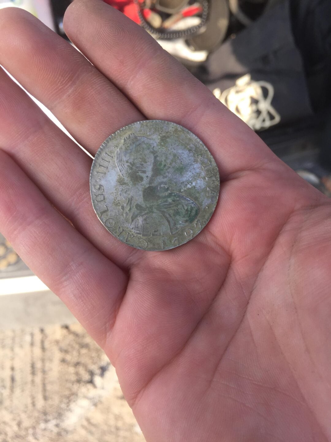 A close-up of a person's hand holding an old, worn coin with a faint image of a person and partially visible text around the edge. The background is slightly blurred.