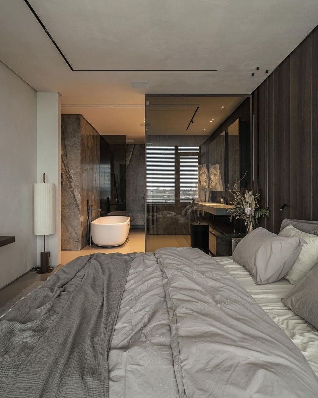 A modern bedroom with a large unmade bed in the foreground, neutral-toned bedding, and floor-to-ceiling glass wall revealing a bathroom with a freestanding bathtub and marble accents.