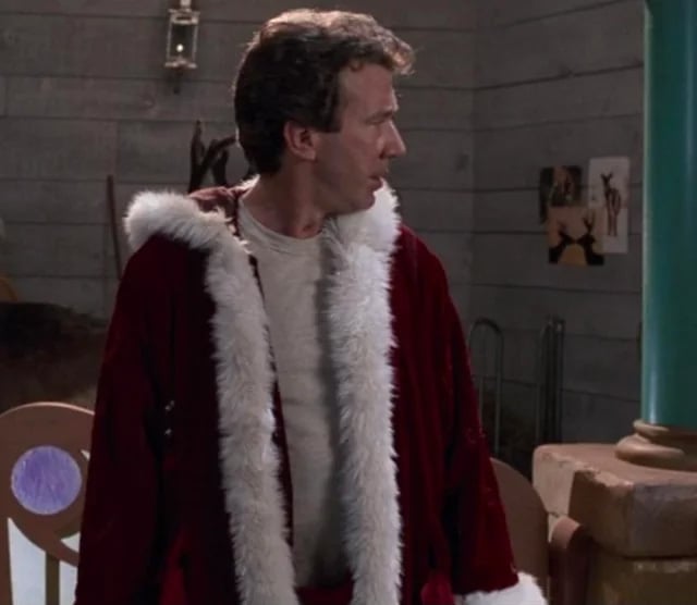 A man wearing a red and white Santa coat over a beige shirt stands indoors, looking to his left. The room has wooden walls, some posters, and a mix of rustic and whimsical decor.