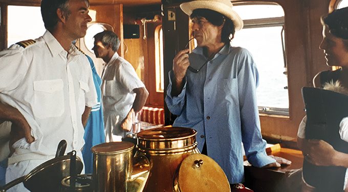 A group of men, some in nautical uniforms and one in a light shirt and hat, stand and talk inside a boat cabin with brass instruments and windows showing the sea outside.