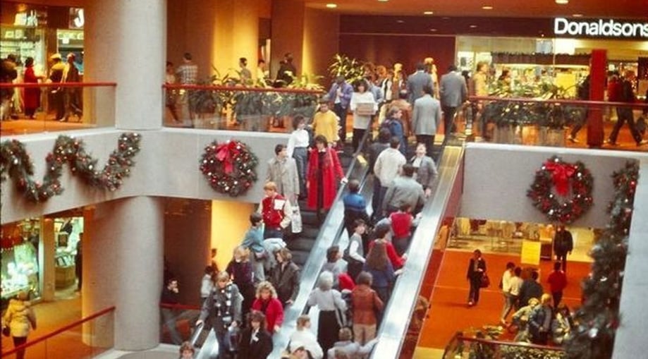 Busy shopping mall decorated for the holidays with wreaths and garlands. Shoppers, some in winter clothes, use escalators and walk through the crowded space. Store signs, including "Donaldson’s," are visible in the background.