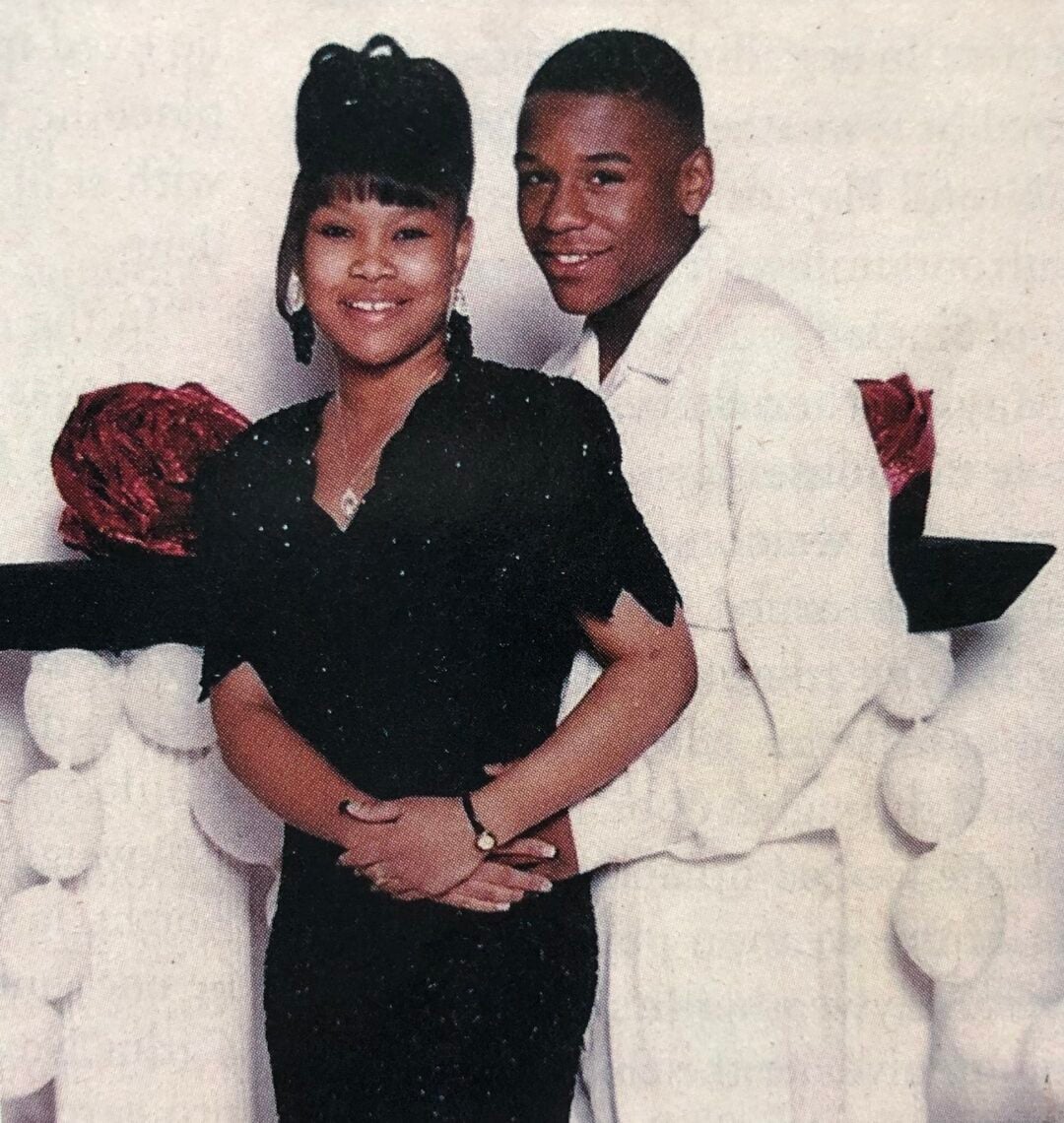 A young couple poses closely together in front of a decorative backdrop. The woman wears a black dress and earrings, while the man stands behind her in a white suit, both smiling softly at the camera.