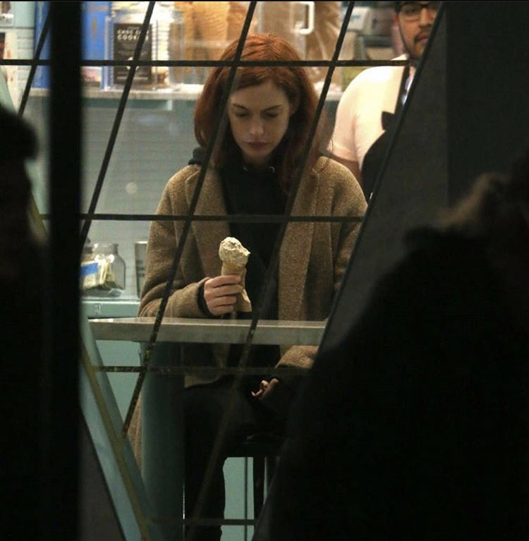 A woman with red hair sits alone at a table in an ice cream shop, looking down at an ice cream cone in her hand. The scene is viewed through a window with diagonal bars.