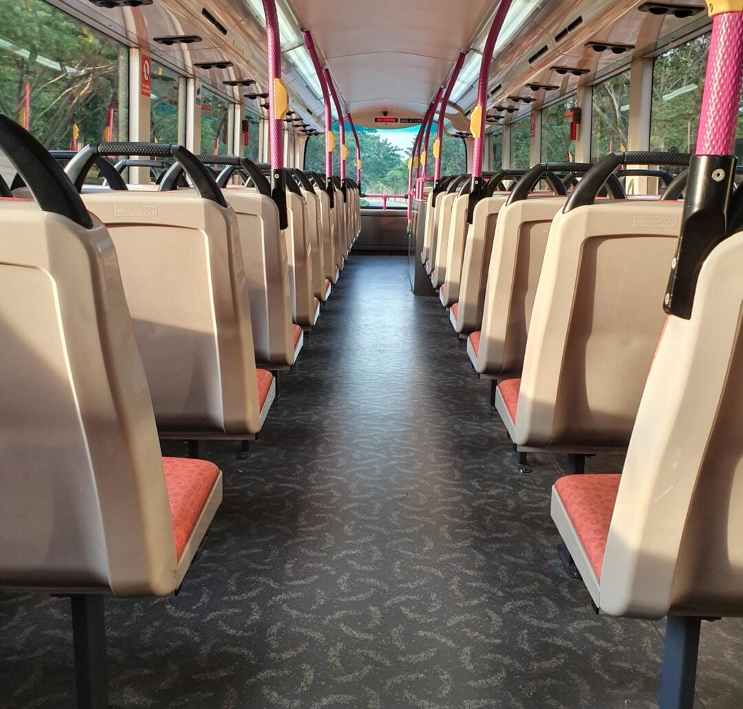 Empty bus interior with rows of pink and beige seats, sunlight streaming in from the windows, and a clear aisle running down the center toward the front of the bus.