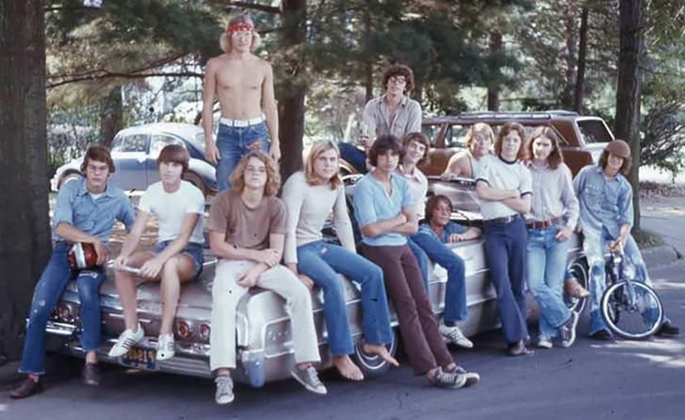 A group of teenagers, mostly boys in casual 1970s clothing, sit and stand on and around a classic white car parked outdoors among trees. One boy is shirtless, and a bicycle rests nearby.