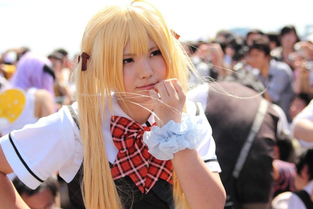 A person in a blonde wig, plaid bowtie, and white school uniform cosplay poses with their hand near their face. A crowd of people and photographers are blurred in the background.
