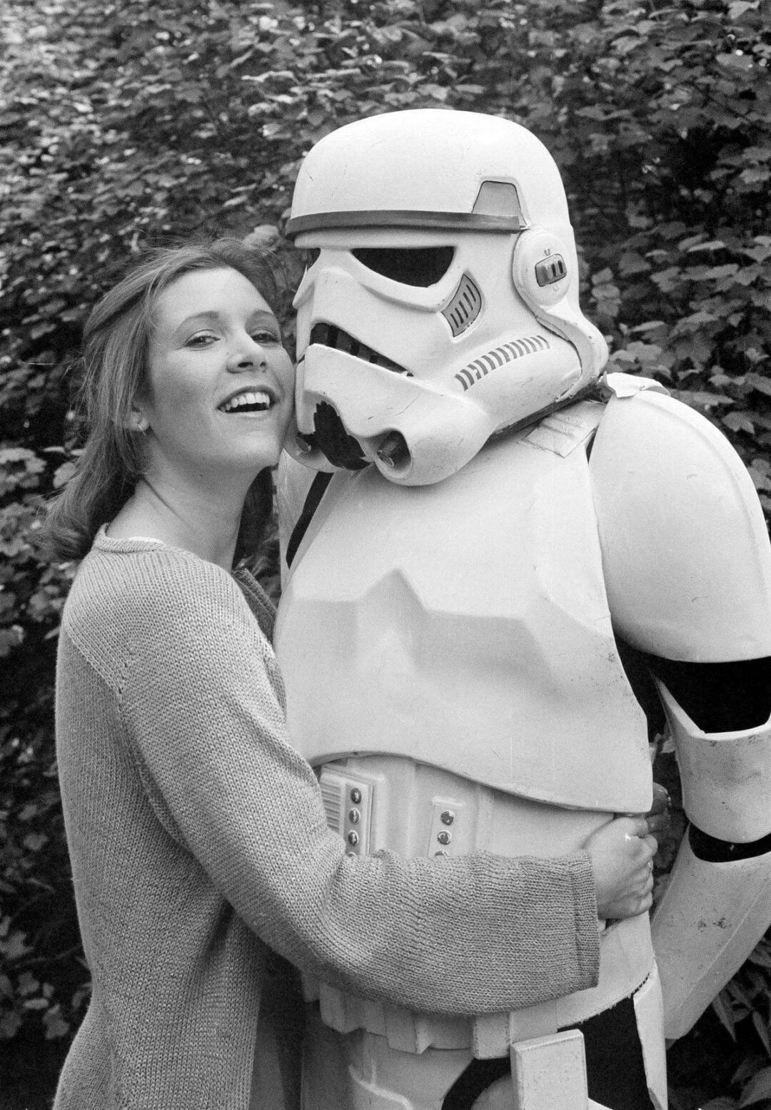 A woman smiling with her arms around a person dressed in a Star Wars stormtrooper costume, standing outdoors in front of leafy bushes.