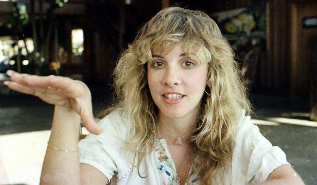 A woman with long, wavy blonde hair and bangs sits indoors, raising her left hand with her fingers spread. She wears a white blouse with embroidery and looks directly at the camera with a relaxed expression.
