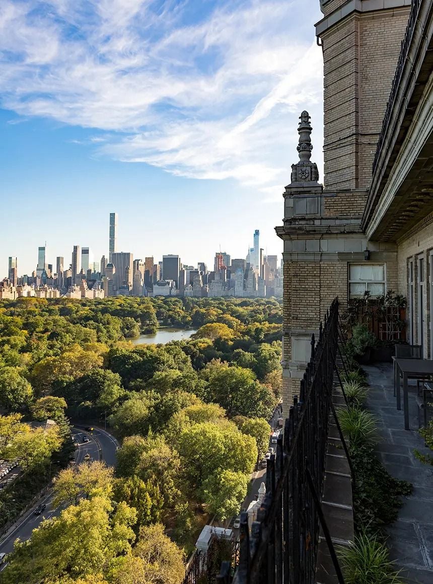 A view from a balcony overlooking Central Park in New York City, with lush green trees below and tall skyscrapers in the distance under a blue sky with wispy clouds.