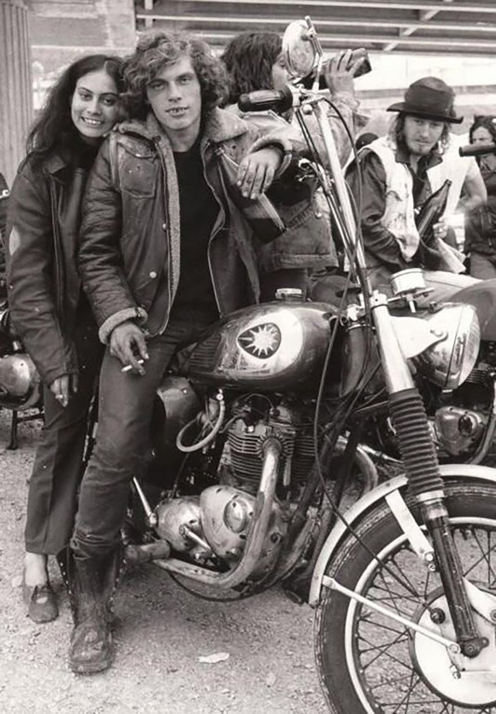 A young man sits on a vintage motorcycle, holding a cigarette, with a smiling woman standing close beside him. Two other people stand in the background, dressed in casual, biker-style clothing. The scene appears to be from the 1970s.