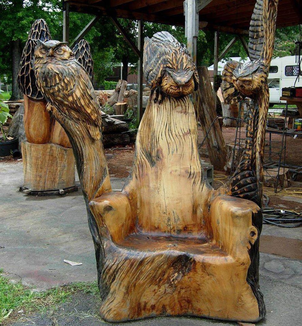 A large wooden chair carved from a single tree trunk, featuring detailed sculptures of three owls with outstretched wings on the backrest, set outdoors in a workshop area.