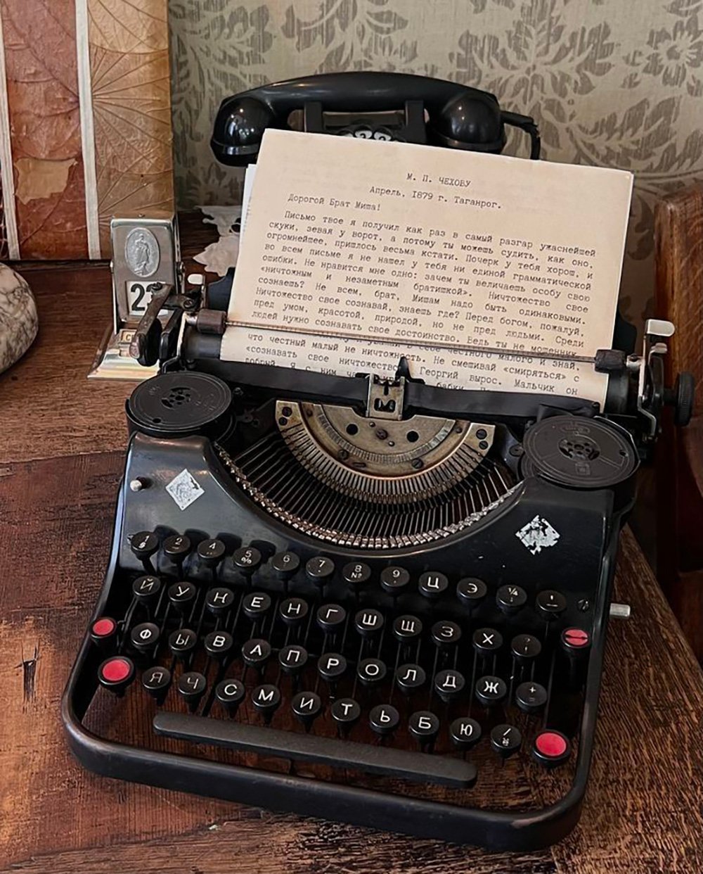 A vintage black typewriter with Russian keys sits on a wooden desk. A typed Russian letter is inserted in the roller. Behind it are an old-fashioned telephone, a calendar, and some decorative papers.
