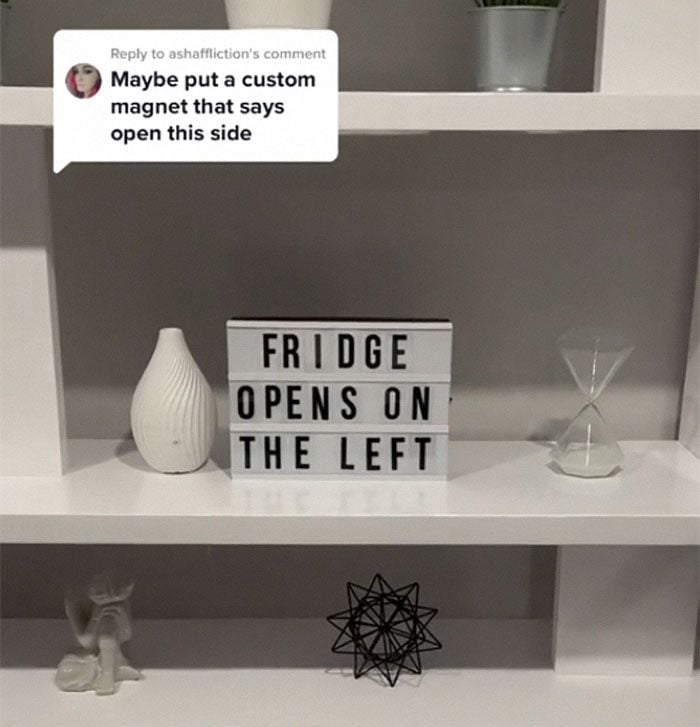 A white shelf displays a sign reading "FRIDGE OPENS ON THE LEFT," an hourglass, a white vase, a geometric black sculpture, and a small white figurine. A comment above suggests using a custom magnet.