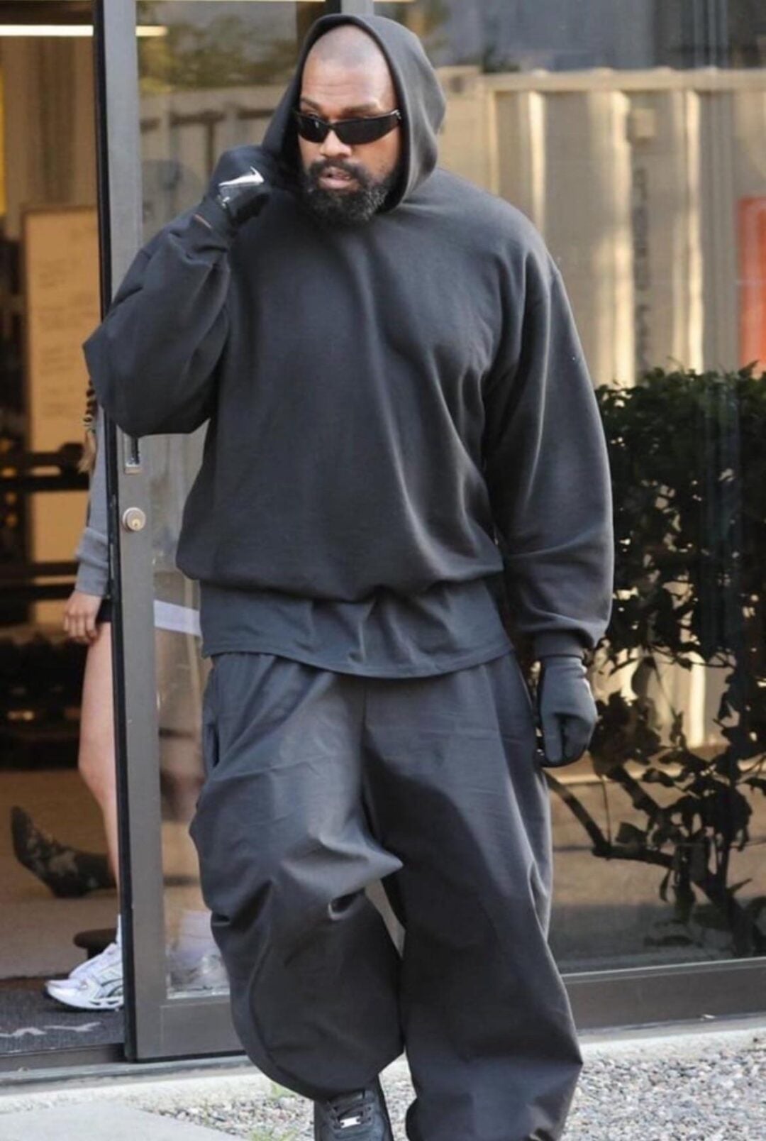 A person wearing a black hooded sweatshirt, baggy black pants, gloves, and sunglasses walks outside near a glass door. Their hand is raised to their face, and they have a beard.