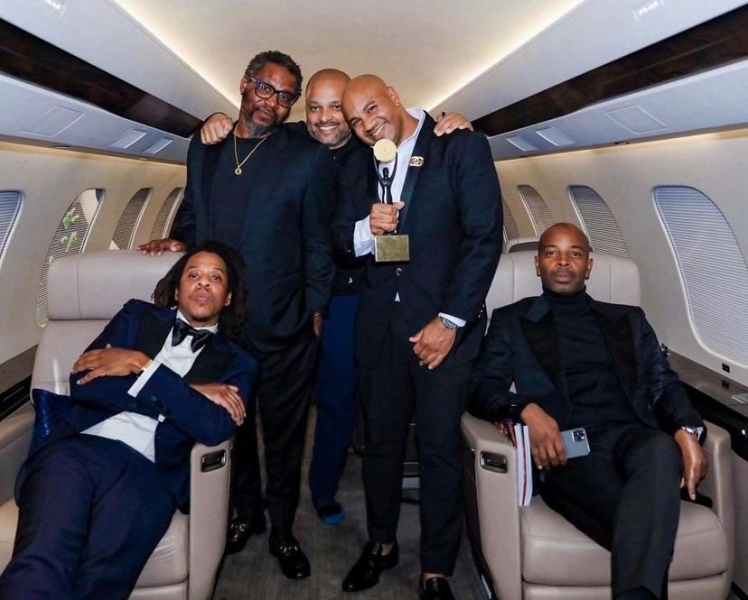 Five men dressed in formal attire pose together inside a private jet. Three stand closely in the aisle, smiling at the camera, while two sit relaxed in leather seats on either side.