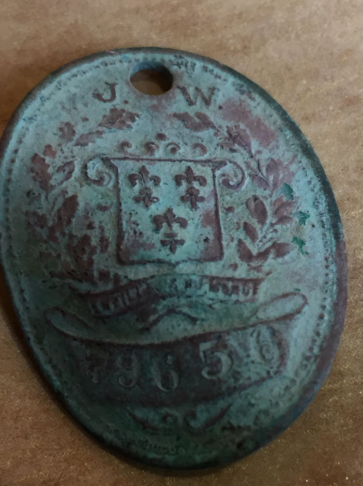 An old, oval-shaped metal tag with a hole at the top, engraved with a shield, floral designs, letters "J W," and the number "9659." The surface appears worn and tarnished with green and reddish-brown patina.