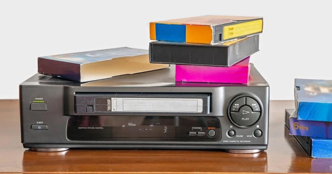 A VCR (videocassette recorder) sits on a wooden surface with several VHS tapes stacked on top of it and nearby. The VCR display is on, and the tapes are in colorful cases.