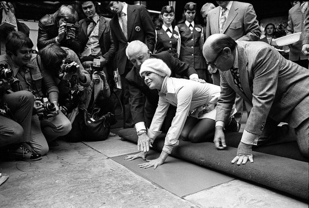 A woman in a white outfit and hat presses her hands into a slab as photographers and onlookers surround her. Two men kneel beside her, assisting with the handprint ceremony. The atmosphere appears bustling and celebratory.