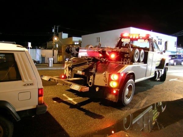 A tow truck with flashing red lights is preparing to tow a white SUV at night on a wet street, with buildings and streetlights visible in the background.