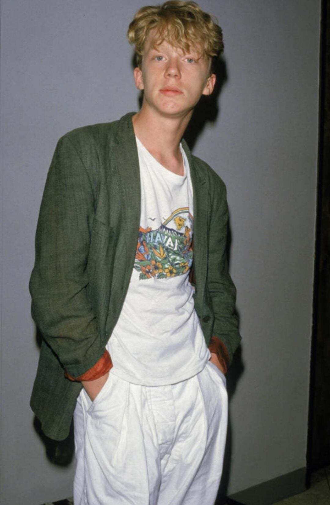 A young person with blond, tousled hair stands with hands in white pants pockets, wearing a green blazer over a printed white T-shirt. The background is plain gray.