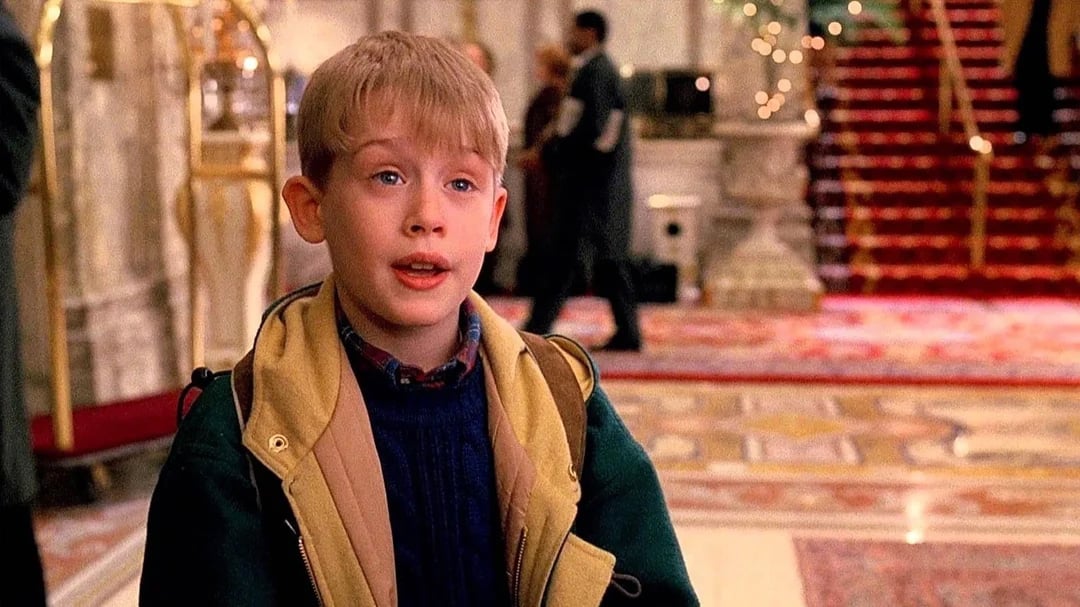 A young blond boy wearing winter clothes stands in the lobby of an elegant hotel, with ornate decor, red carpeted stairs, and blurred people in the background.