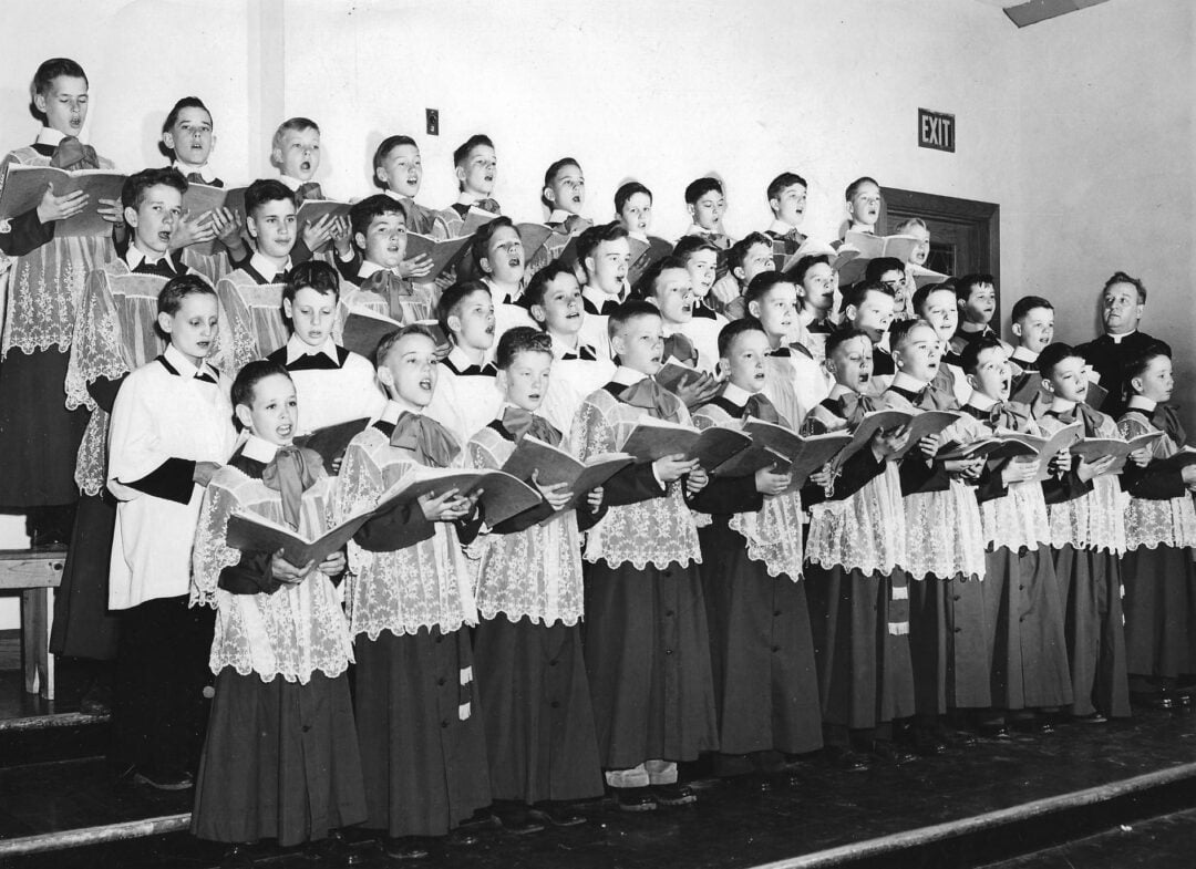 A black and white photo shows a large group of boys in choir robes singing from sheet music, standing on risers in a room with plain walls and an exit sign above a door in the background.