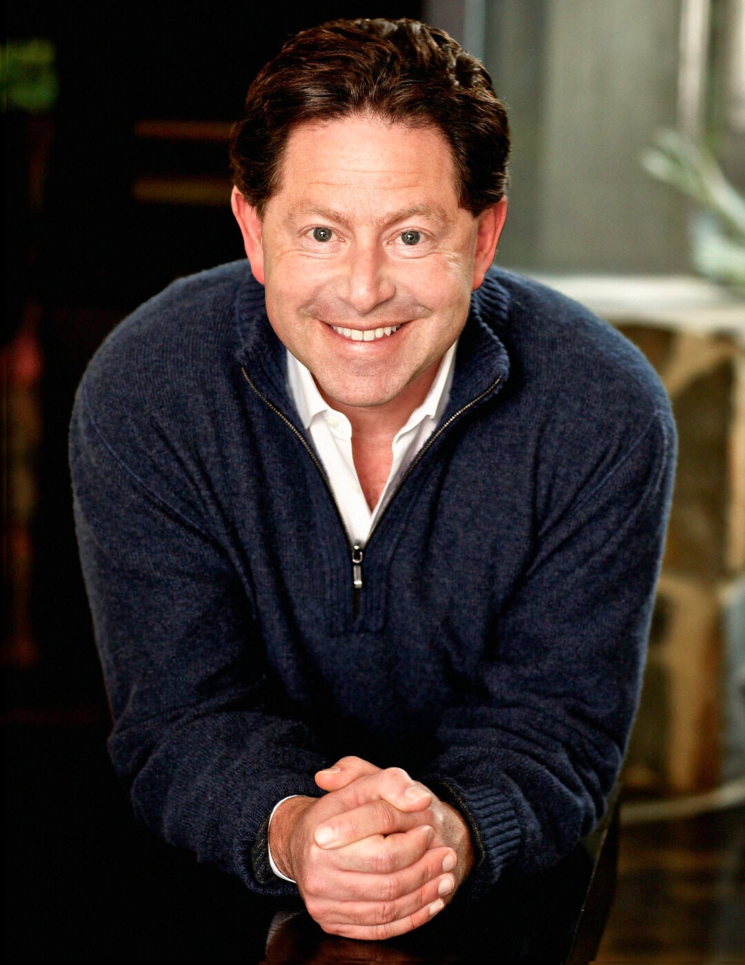 A middle-aged man with curly brown hair wearing a navy blue sweater sits with his hands clasped, looking directly at the camera against a dark background.