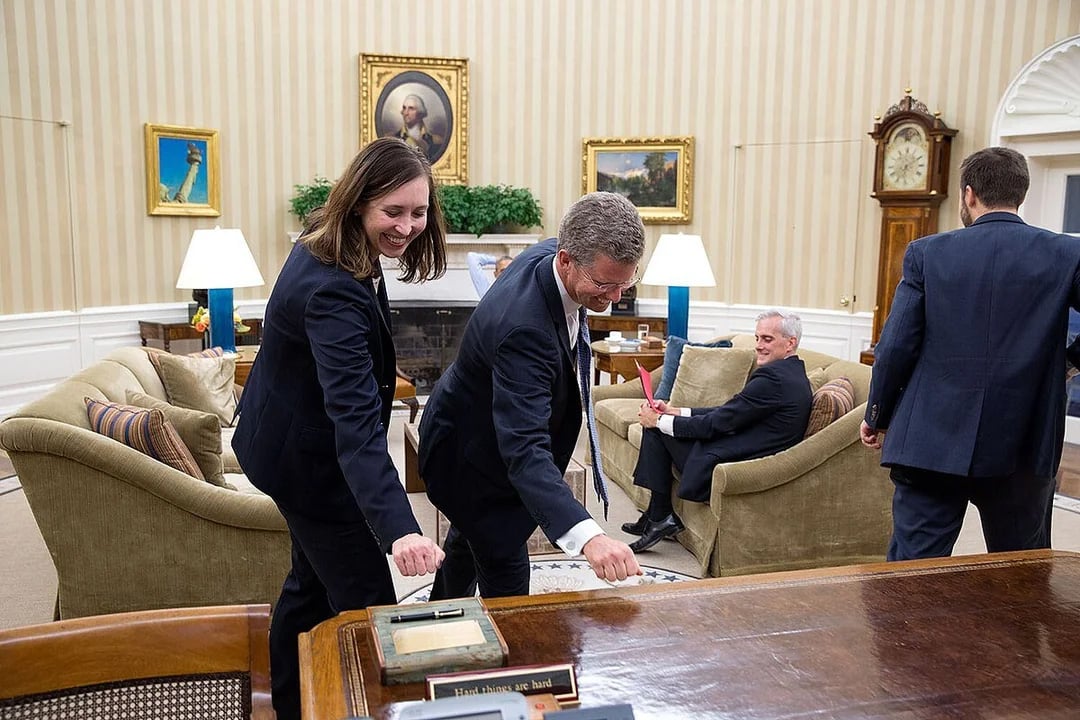 Two people in suits lean forward, appearing to fist bump, beside a large wooden desk in the Oval Office. A man sits on a couch holding papers, while another person walks away. The room is decorated with paintings and lamps.